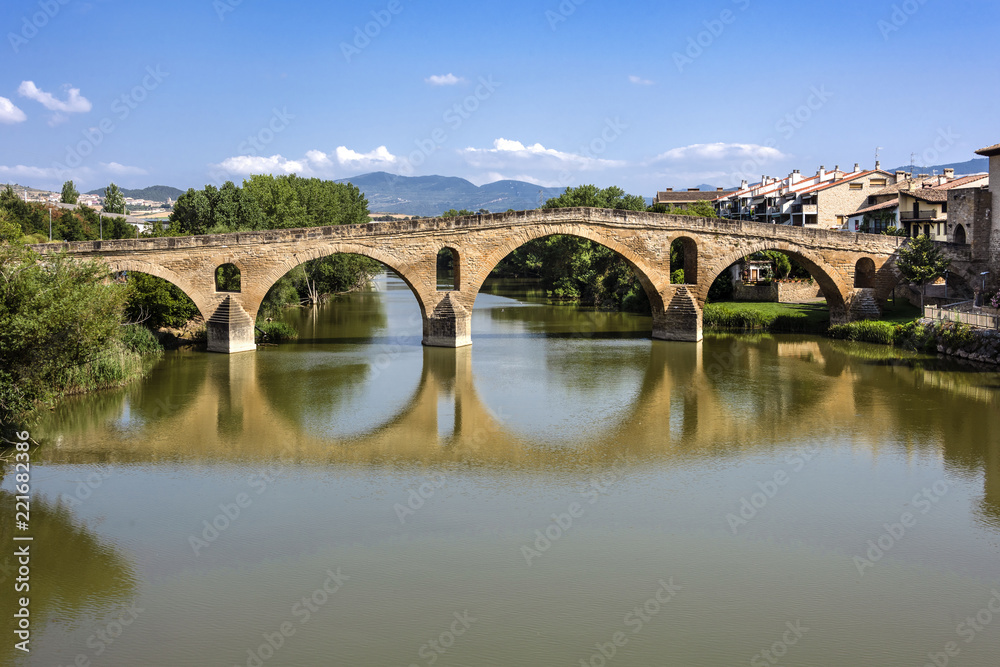 Spain, Puente La Reina, Gares: Panorama view of famous romanesque bridge over river Arga with skyline of Spanish small town literally named 'Bridge of the Queen', green riverside and blue sky.