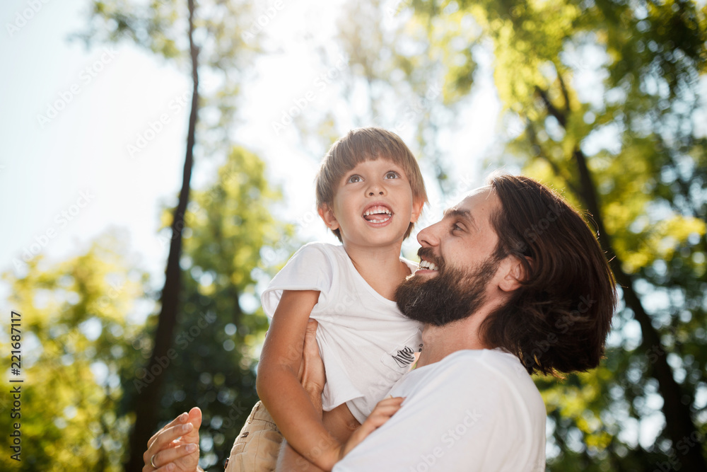 Fototapeta premium Handsome dark-haired father with beard dressed in the white t-shirt is holding in the arms his little son, watching at him and smiling in the park.