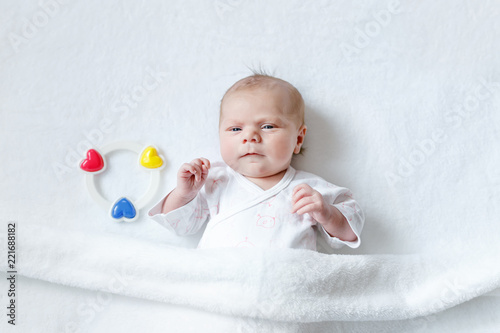 Cute adorable newborn baby playing with colorful rattle toy. in white bed at home. New born child, little girl looking surprised at the camera. Family, new life, childhood, beginning concept.