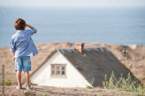 lonely little boy looking far away towards sea horizon near old house