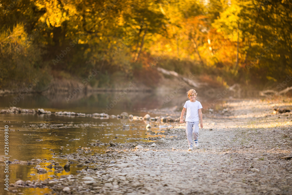 Little blonde boy in green glasses playing on the riverside. Autumn in the yellow forest Little blonde boy in green glasses playing on the riverside. Autumn in the yellow forest