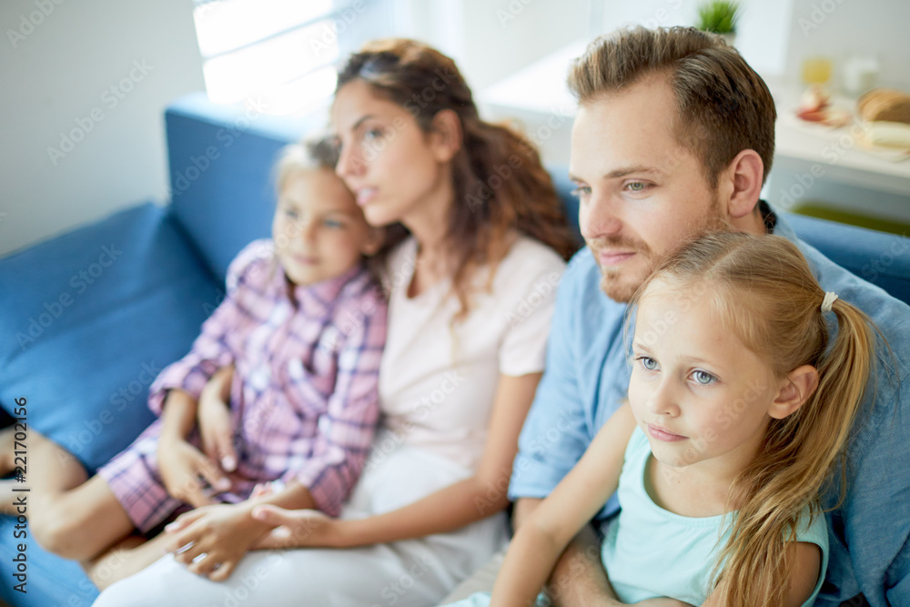 Young dad and mom holding little daughters while all sitting on sofa in front of tv set and watching movie at leisure