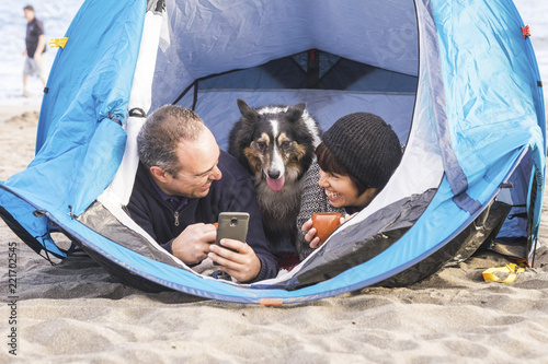 couple looking at the smart phone and have fun inside a tent in free camping on the beach Dog border collie behind them looking at the camera. bright colors and alternative vacation family concept