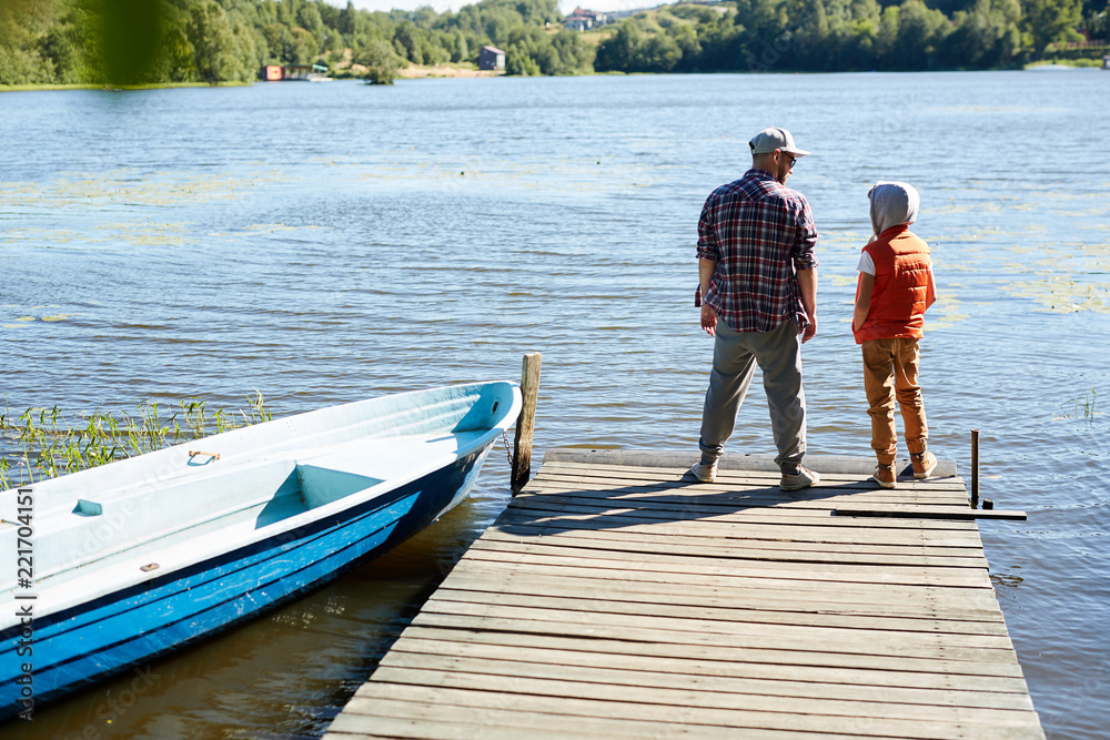 Rear view of dad and son standing on wooden pontoon in front of waterside with boat near by