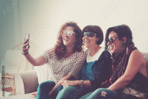 three caucasian young beautiful females doing a picture in selfie style with a mobile phone at home sit down on the sofa. friendship and connected with friends with wifi technology.. home interior photo