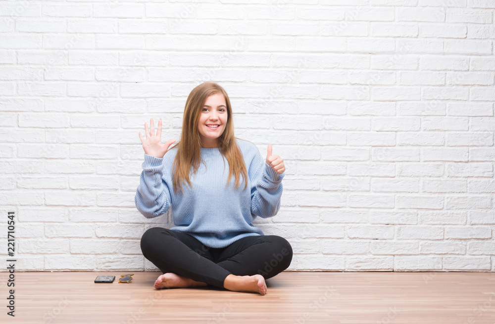 Young adult woman sitting on the floor in autumn over white brick wall showing and pointing up with fingers number six while smiling confident and happy. Young adult woman sitting on the floor in autumn over white brick wall showing and pointing up with fingers number six while smiling confident and happy.