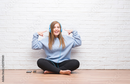 Young adult woman over white brick wall listening music wearing headphones very happy pointing with hand and finger