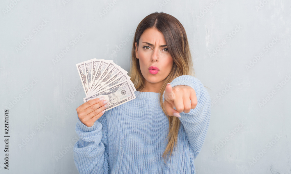 Young adult woman over grey grunge wall holding dollars pointing with finger to the camera and to you, hand sign, positive and confident gesture from the front Young adult woman over grey grunge wall holding dollars pointing with finger to the camera and to you, hand sign, positive and confident gesture from the front