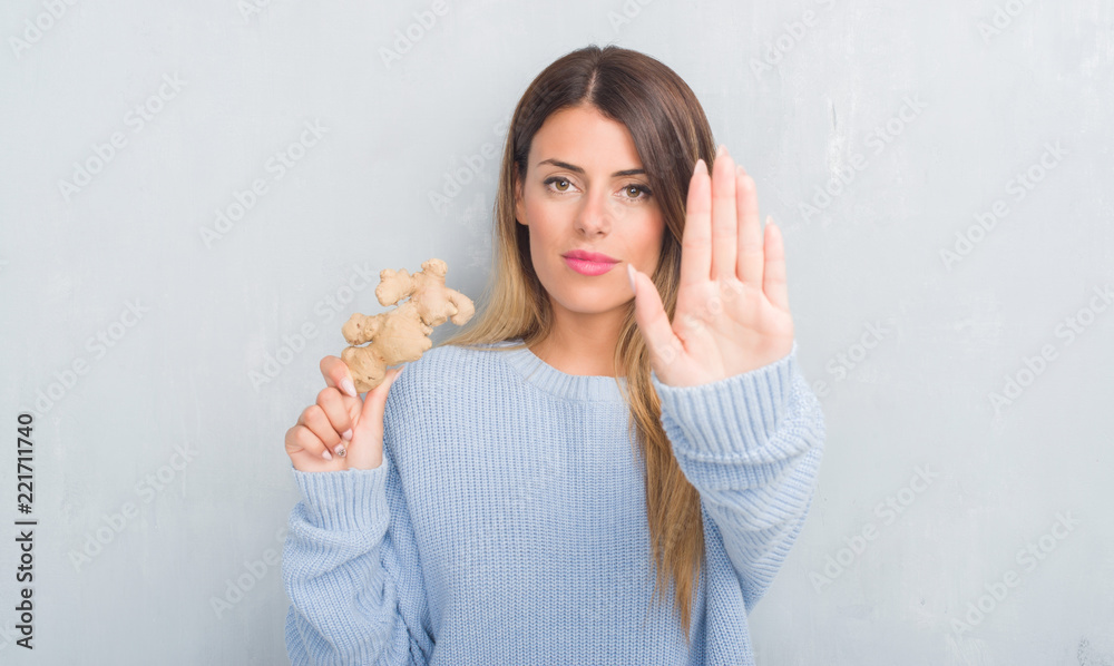 Young adult woman over grey grunge wall holding fresh ginger vegatable with open hand doing stop sign with serious and confident expression, defense gesture Young adult woman over grey grunge wall holding fresh ginger vegatable with open hand doing stop sign with serious and confident expression, defense gesture