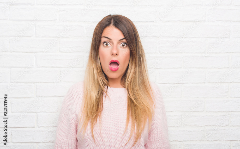 Young adult woman over white brick wall wearing winter outfit at home afraid and shocked with surprise expression, fear and excited face. Young adult woman over white brick wall wearing winter outfit at home afraid and shocked with surprise expression, fear and excited face.