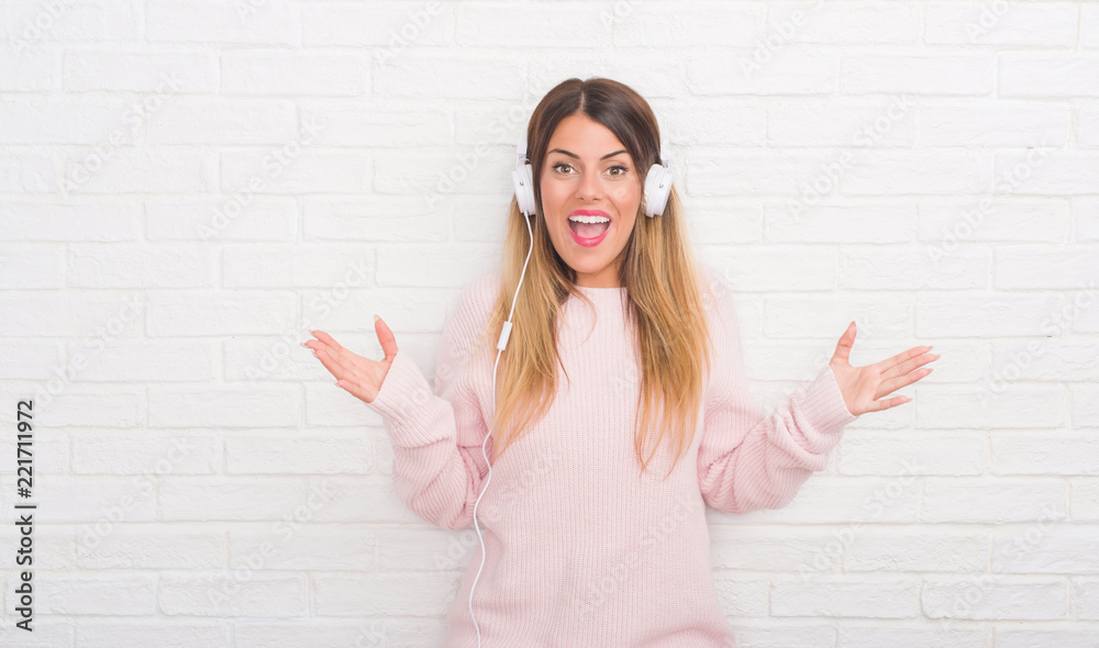 Young adult woman over white brick wall wearing headphones listening to music and dancing very happy and excited, winner expression celebrating victory screaming with big smile and raised hands Young adult woman over white brick wall wearing headphones listening to music and dancing very happy and excited, winner expression celebrating victory screaming with big smile and raised hands