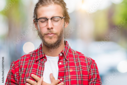 Young handsome man with long hair wearing glasses over isolated background smiling with hands on chest with closed eyes and grateful gesture on face. Health concept.
