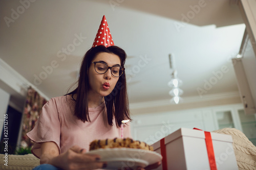 A girl in a cap alone with a cake with candles sitting on the sofa in the room.