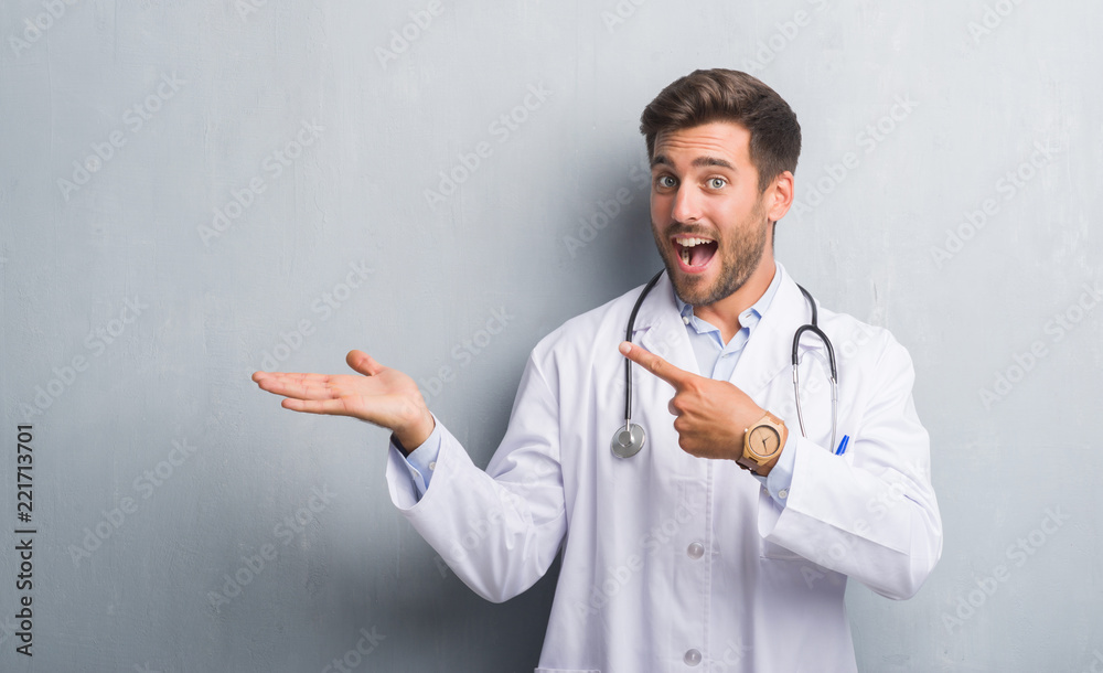 Handsome young doctor man over grey grunge wall amazed and smiling to the camera while presenting with hand and pointing with finger. Handsome young doctor man over grey grunge wall amazed and smiling to the camera while presenting with hand and pointing with finger.