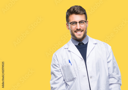 Young handsome man wearing doctor, scientis coat over isolated background looking away to side with smile on face, natural expression. Laughing confident.