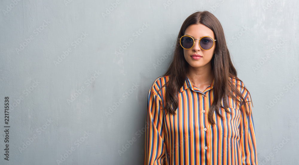 Young brunette woman over grunge grey wall wearing retro sunglasses and outfit with a confident expression on smart face thinking serious
