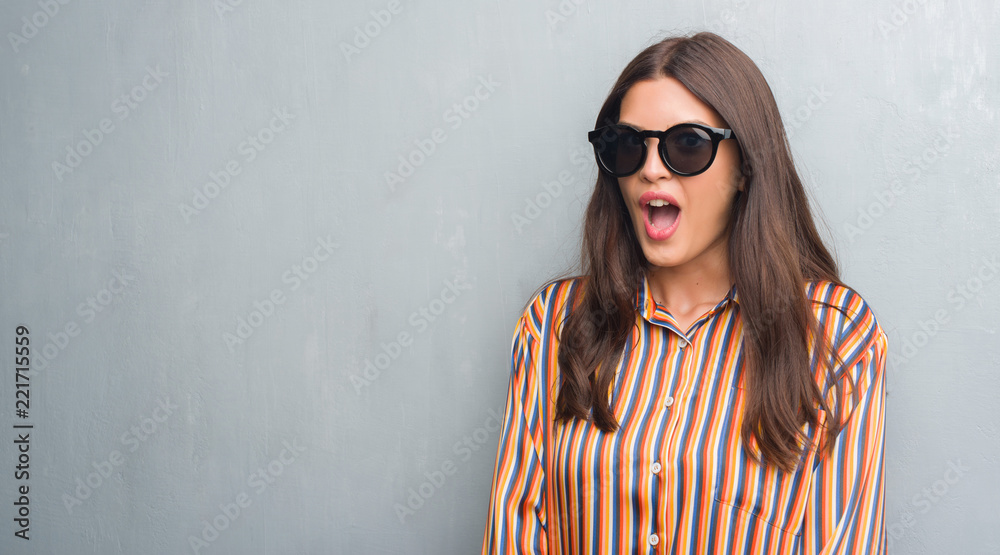 Young brunette woman over grunge grey wall wearing big sunglasses scared in shock with a surprise face, afraid and excited with fear expression Young brunette woman over grunge grey wall wearing big sunglasses scared in shock with a surprise face, afraid and excited with fear expression