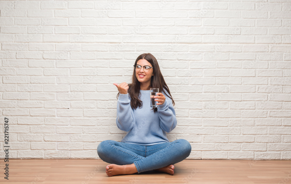 Young brunette woman sitting on the floor drinking glass of water pointing and showing with thumb up to the side with happy face smiling Young brunette woman sitting on the floor drinking glass of water pointing and showing with thumb up to the side with happy face smiling