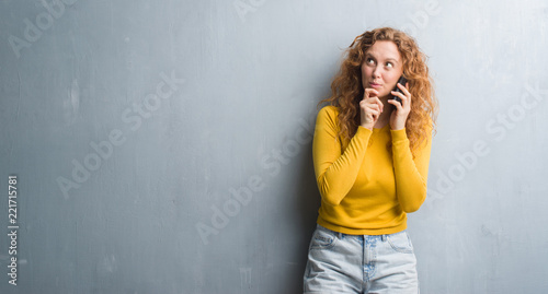 Young redhead woman over grey grunge wall talking on the phone serious face thinking about question, very confused idea