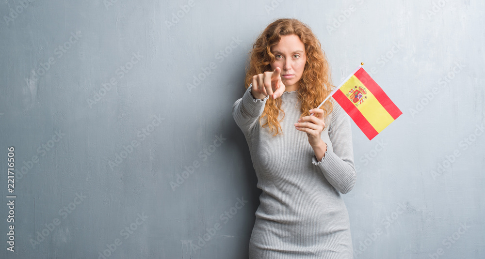 Young redhead woman over grey grunge wall holding flag of Spain pointing with finger to the camera and to you, hand sign, positive and confident gesture from the front Young redhead woman over grey grunge wall holding flag of Spain pointing with finger to the camera and to you, hand sign, positive and confident gesture from the front