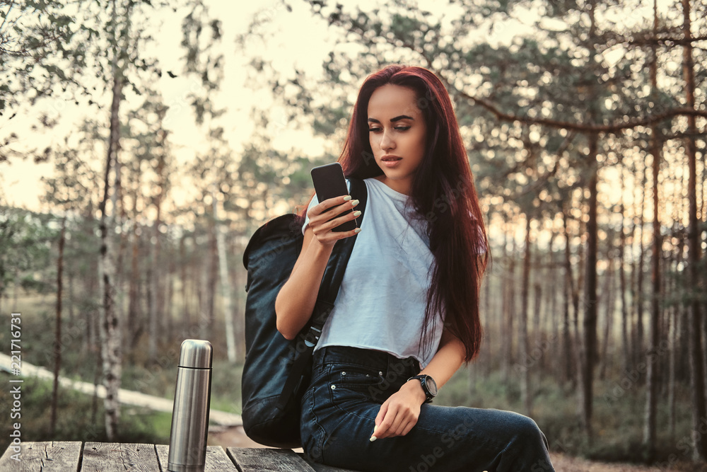 Sensual tourist girl sitting on a wooden table and using a smartphone, having a break in beautiful autumn forest at sunset. Sensual tourist girl sitting on a wooden table and using a smartphone, having a break in beautiful autumn forest at sunset.