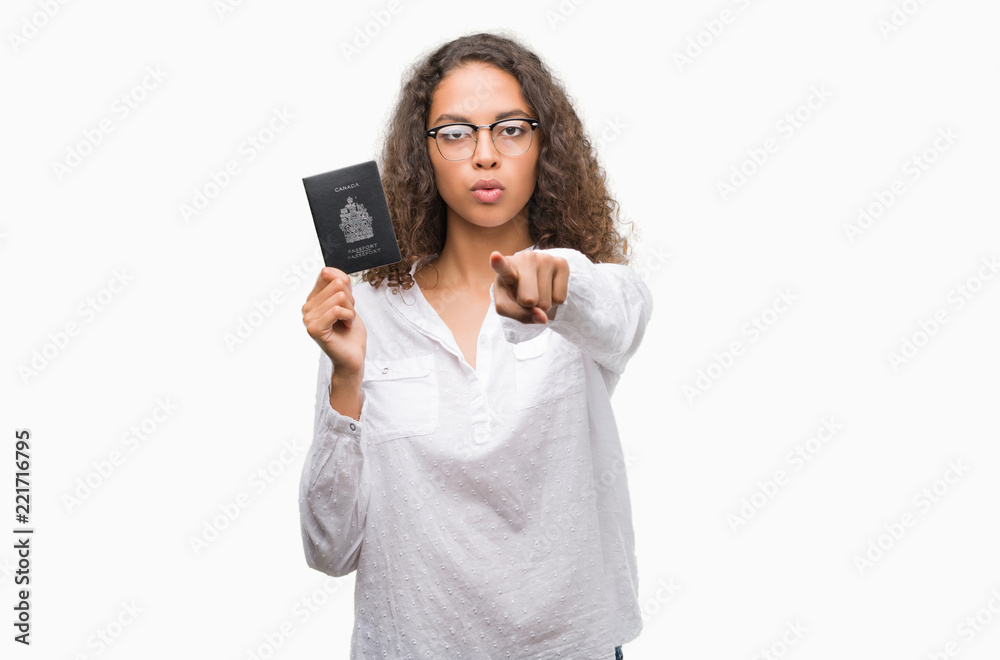 Young hispanic woman holding passport of Canada pointing with finger to the camera and to you, hand sign, positive and confident gesture from the front Young hispanic woman holding passport of Canada pointing with finger to the camera and to you, hand sign, positive and confident gesture from the front