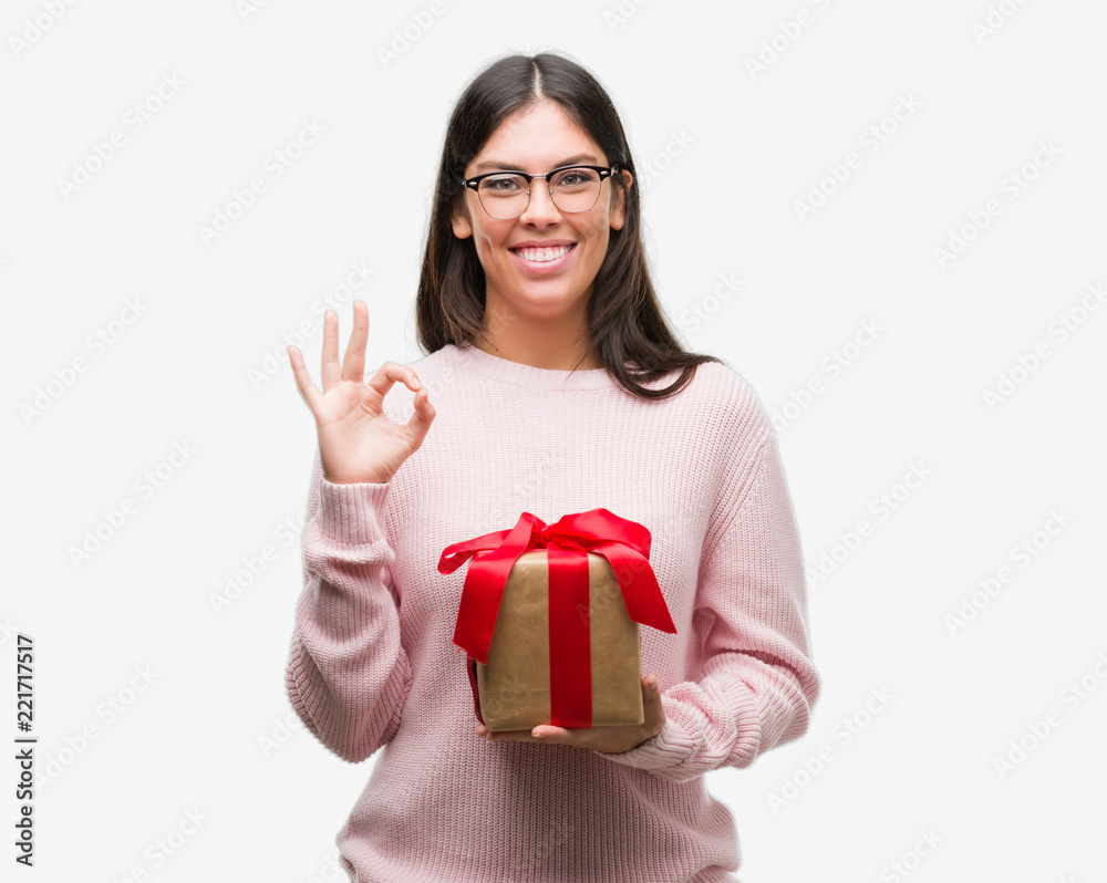 Young hispanic woman holding a gift doing ok sign with fingers, excellent symbol Young hispanic woman holding a gift doing ok sign with fingers, excellent symbol