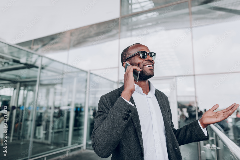 Waist up portrait of happy guy in sunglasses is having call, while standing at the entrance of airport Waist up portrait of happy guy in sunglasses is having call, while standing at the entrance of airport