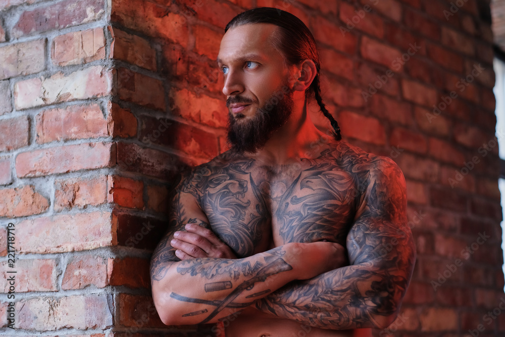 Stylish bearded male with the muscular and tattooed body leaning on a wall with crossed arms and looking out the window. Stylish bearded male with the muscular and tattooed body leaning on a wall with crossed arms and looking out the window.