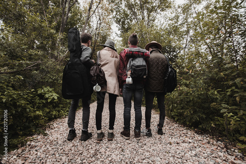 Back view of group of teenagers in warm clothes with guitar and backpacks standing in line on rocky road in forest