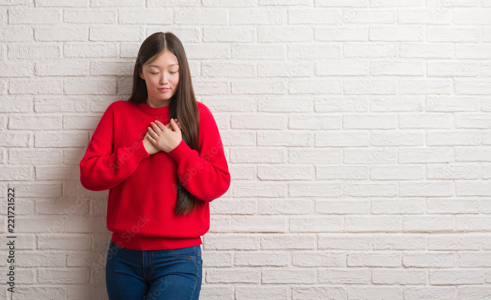Young Chinese woman over brick wall smiling with hands on chest with closed eyes and grateful gesture on face. Health concept.