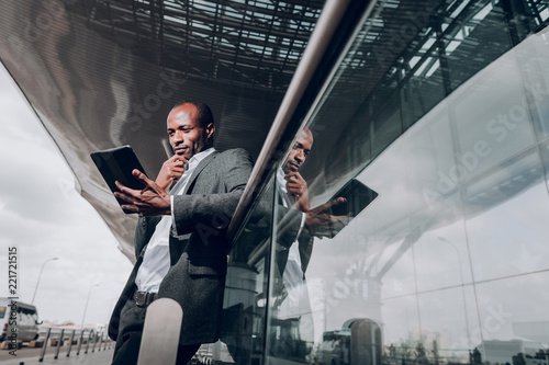 Lets think. Low angle portrait of young male leaning on glass fence at the airport. Man is holding tablet with pensive look