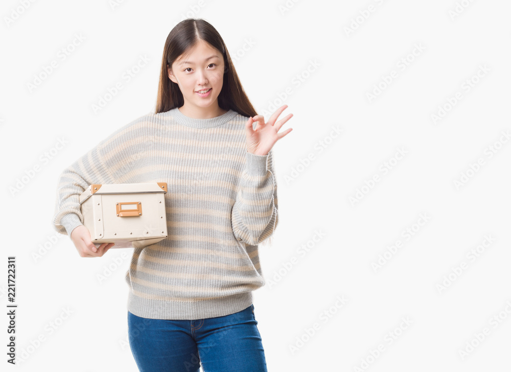 Young Chinese woman over isolated background holding a box doing ok sign with fingers, excellent symbol Young Chinese woman over isolated background holding a box doing ok sign with fingers, excellent symbol