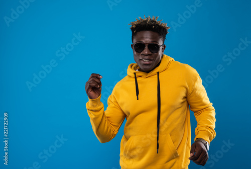 Having good mood. Waist up portrait of young man moving to the rhythm of music. He is wearing yellow sweatshirt and looking at camera with smile. Copy space in left side