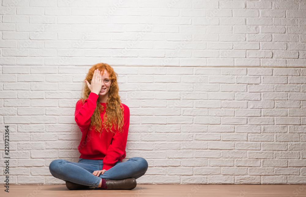 Young redhead woman sitting over brick wall covering one eye with hand with confident smile on face and surprise emotion. Young redhead woman sitting over brick wall covering one eye with hand with confident smile on face and surprise emotion.