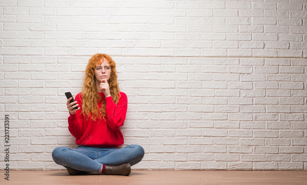 Young redhead woman sitting over brick wall talking on the phone serious face thinking about question, very confused idea