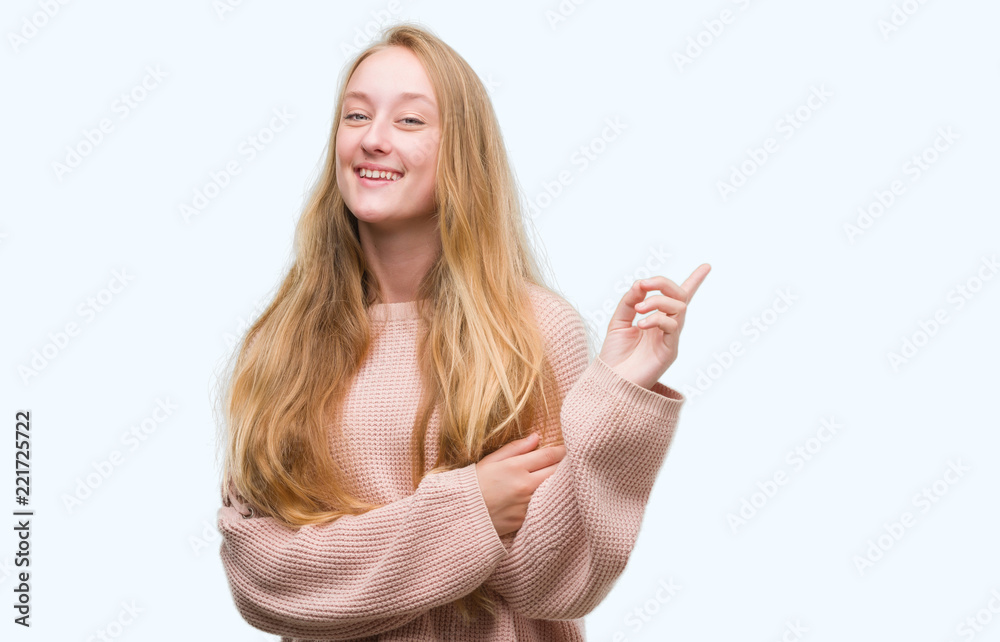 Blonde teenager woman wearing pink sweater with a big smile on face, pointing with hand and finger to the side looking at the camera. Blonde teenager woman wearing pink sweater with a big smile on face, pointing with hand and finger to the side looking at the camera.