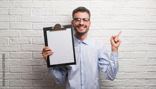 Young adult business man over brick wall holding clipboard very happy pointing with hand and finger to the side