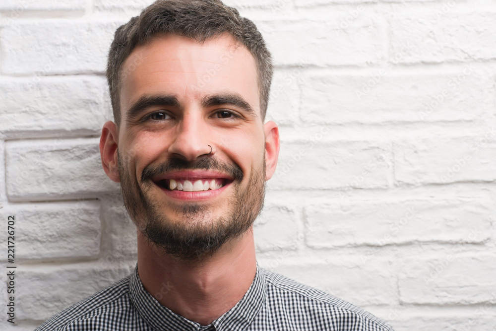 Young adult man standing over white brick wall with a happy face standing and smiling with a confident smile showing teeth Young adult man standing over white brick wall with a happy face standing and smiling with a confident smile showing teeth