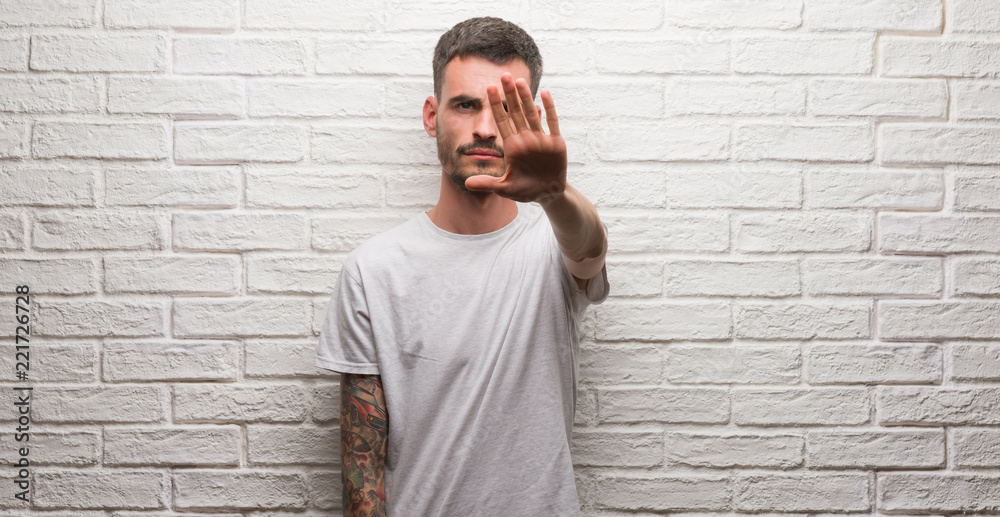 Young adult man standing over white brick wall with open hand doing stop sign with serious and confident expression, defense gesture Young adult man standing over white brick wall with open hand doing stop sign with serious and confident expression, defense gesture