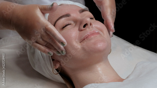 Skin rejuvenation in the cosmetology center. A girl with closed eyes smiles at a procedure in a beauty salon. Hands of the cosmetologist in gloves put a cream on a woman's face.