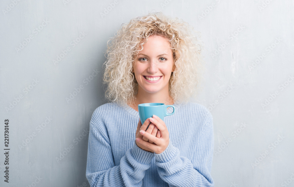 Young blonde woman over grunge grey wall driking a cup of coffee with a happy face standing and smiling with a confident smile showing teeth Young blonde woman over grunge grey wall driking a cup of coffee with a happy face standing and smiling with a confident smile showing teeth
