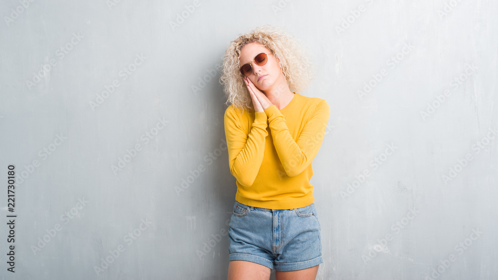 Young blonde woman with curly hair over grunge grey background sleeping tired dreaming and posing with hands together while smiling with closed eyes. Young blonde woman with curly hair over grunge grey background sleeping tired dreaming and posing with hands together while smiling with closed eyes.