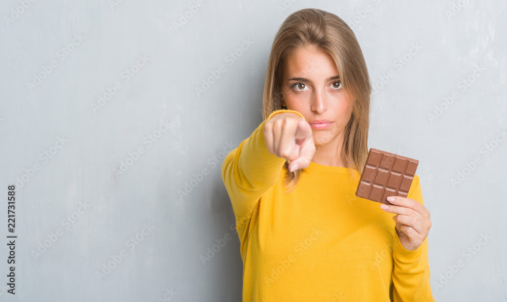 Beautiful young woman over grunge grey wall eating chocolate bar pointing with finger to the camera and to you, hand sign, positive and confident gesture from the front Beautiful young woman over grunge grey wall eating chocolate bar pointing with finger to the camera and to you, hand sign, positive and confident gesture from the front
