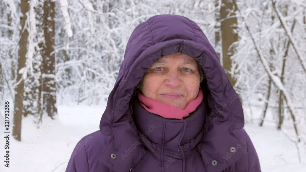 Close up of attractive senior woman in winter jacket with hood walking forward, looking into camera and smiling in winter forest covered with snow. Shallow depth of field, 4K Close up of attractive senior woman in winter jacket with hood walking forward, looking into camera and smiling in winter forest covered with snow. Shallow depth of field, 4K