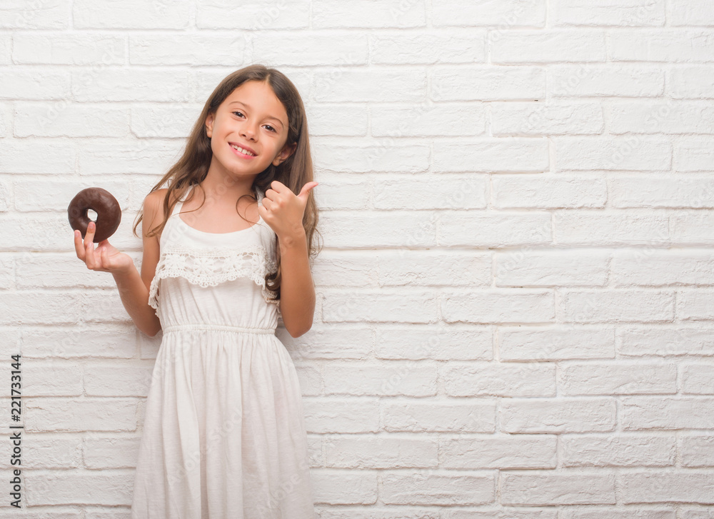Young hispanic kid over white brick wall eating chocolate donut happy with big smile doing ok sign, thumb up with fingers, excellent sign Young hispanic kid over white brick wall eating chocolate donut happy with big smile doing ok sign, thumb up with fingers, excellent sign
