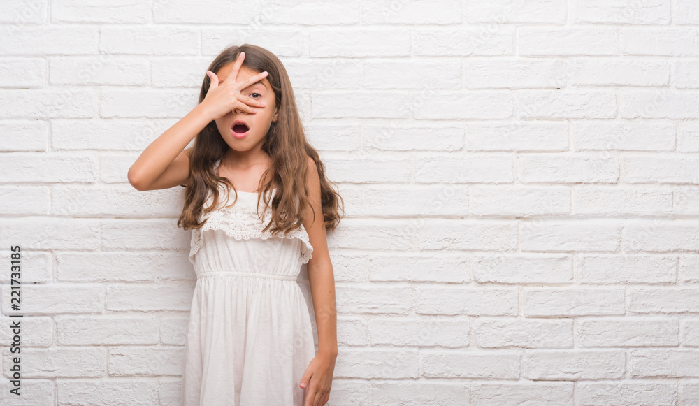 Young hispanic kid over white brick wall peeking in shock covering face and eyes with hand, looking through fingers with embarrassed expression. Young hispanic kid over white brick wall peeking in shock covering face and eyes with hand, looking through fingers with embarrassed expression.