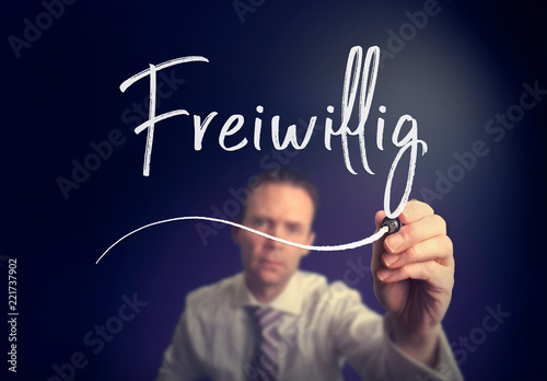 A businessman writing a Voluntary "Freiwillig" concept in German with a white pen on a clear screen.