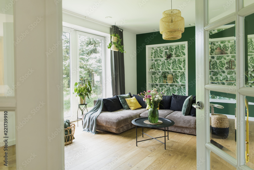 Naklejka premium Stylish interior with sofa, colors pillows, flowers and wooden floor. Sunny and bright living room with big window and design commode.. 