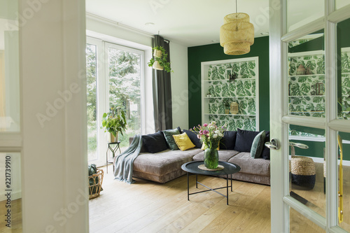 Fototapeta Naklejka Na Ścianę i Meble -  Stylish interior with sofa, colors pillows, flowers and wooden floor. Sunny and bright living room with big window and design commode.. 